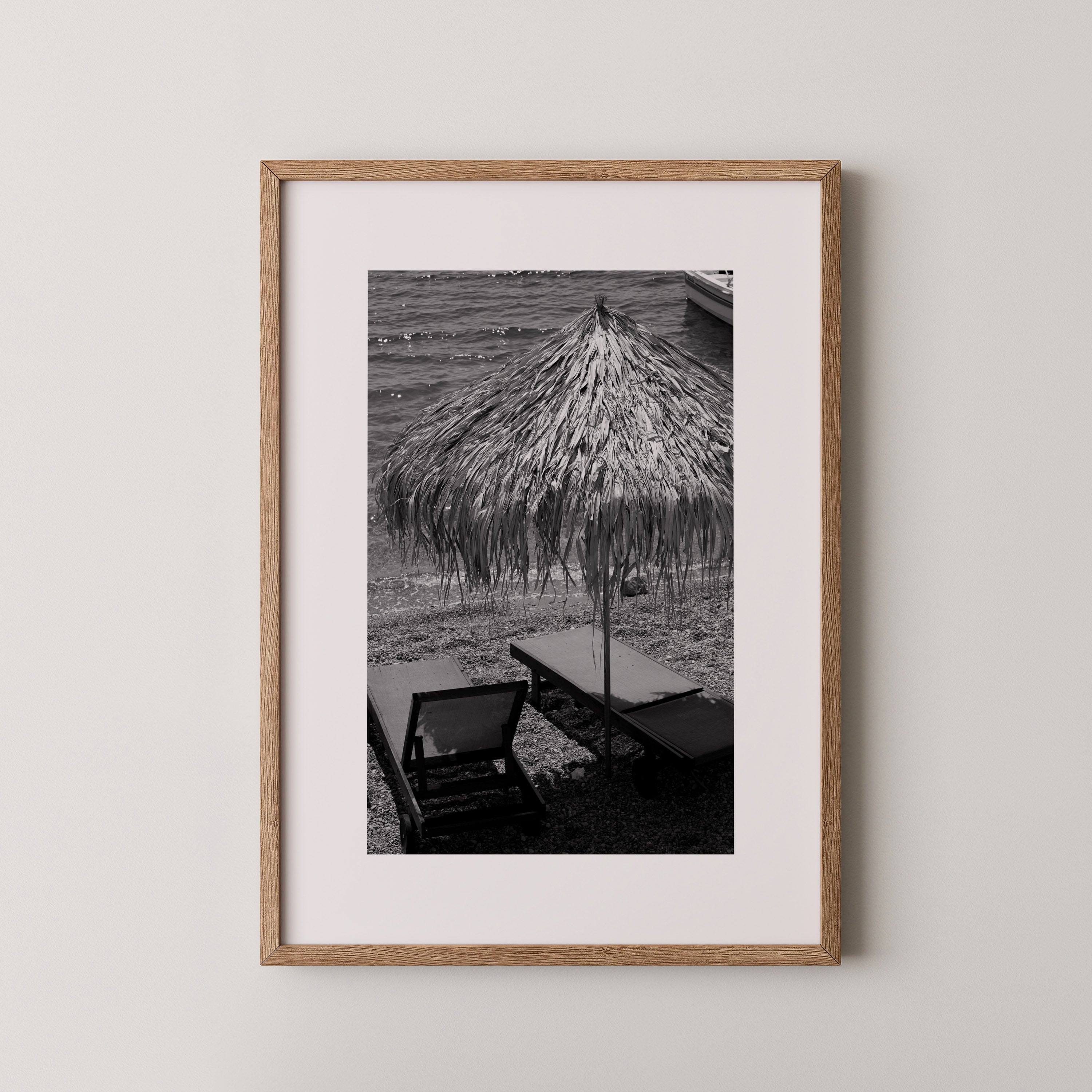 Seaside Pause by Daniel Barlot – black and white Fine Art photograph of a quiet Mediterranean beach with a traditional straw umbrella and empty sun loungers, captured in Greece and framed in natural oak.